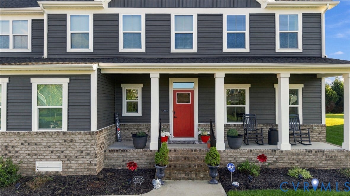 Two-story residence featuring grey siding, a red entry door, white trim, and a covered porch with brick detailing
