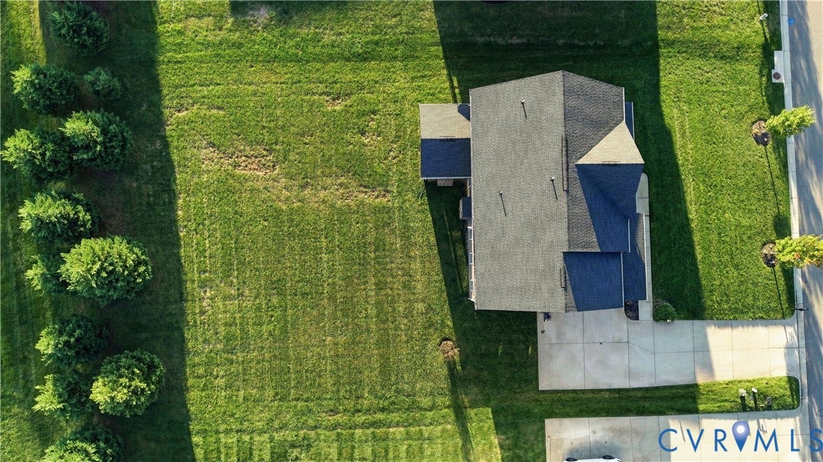 Aerial property view featuring a dark shingled roof, expansive green lawn, mature perimeter trees, and a concrete driveway