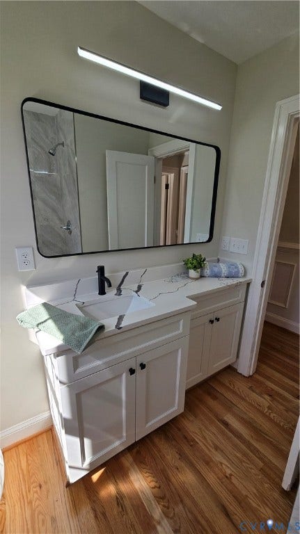 White shaker-style cabinetry with matte black hardware, white countertop with an integrated rectangular sink, wall-mounted rectangular mirror with a black frame, contemporary vanity lighting fixture, and wood-finish flooring