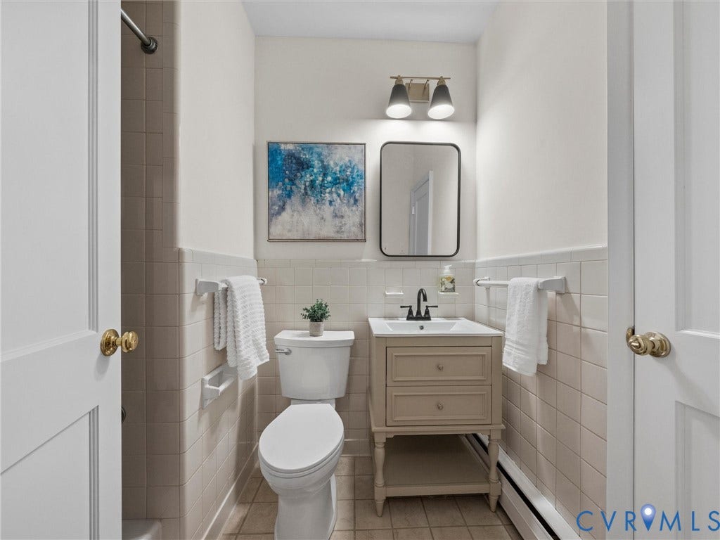 6208 Tapoan Place, Three Chopt, Richmond, VA 23226 White paneled doors with brass hardware, tile wainscoting, built-in vanity with under-sink storage, black framed mirror, and a wall-mounted light fixture