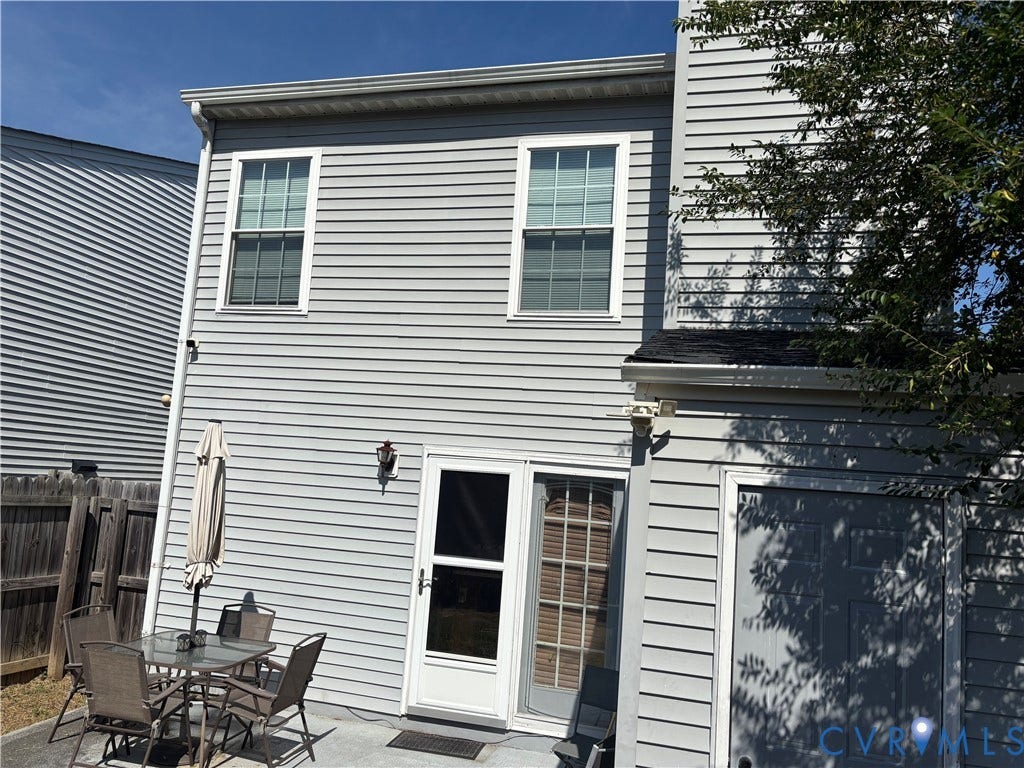 Grey vinyl siding exterior with two upper-level windows, a ground-level patio, a white door with an attached screen door, and a built-in exterior light fixture