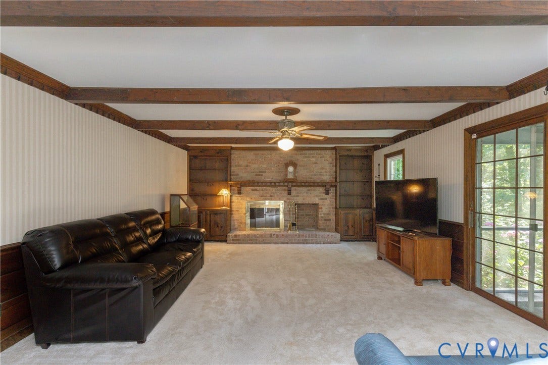 Exposed wood beam ceiling, brick fireplace with hearth, built-in shelving, wood paneled walls, and sliding glass door