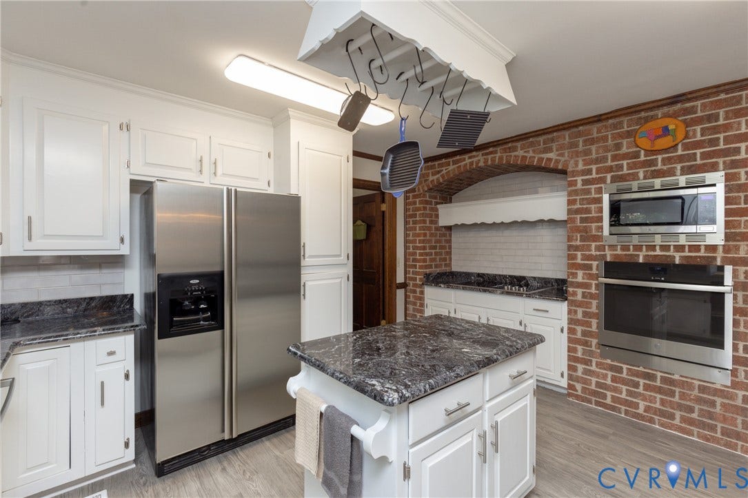 White shaker-style cabinetry, dark stone countertops, stainless steel appliances, brick-finish wall, and wood-finish flooring