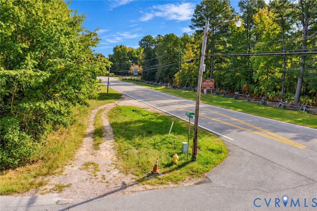 Paved road frontage with a gravel access path, bordered by mature trees and a split-rail fence