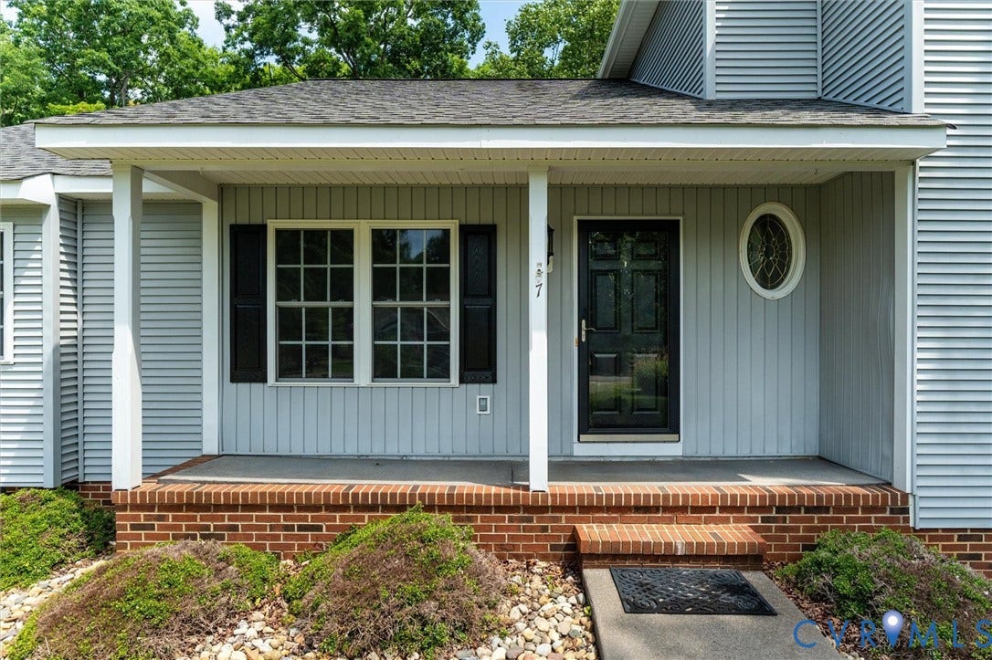 Covered porch entrance with brick foundation, black front door, and an oval window accent