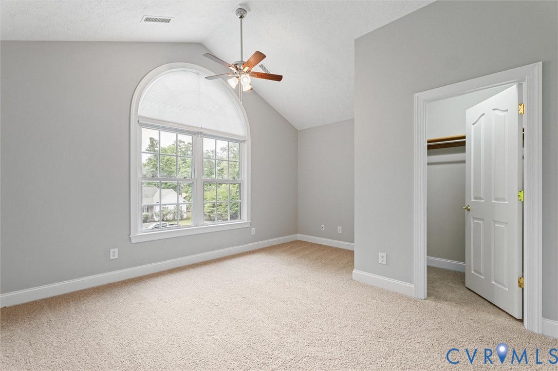 Arched transom window above a multi-pane window, vaulted ceiling, ceiling fan with light fixture, light-colored carpet, and an open closet with built-in shelving