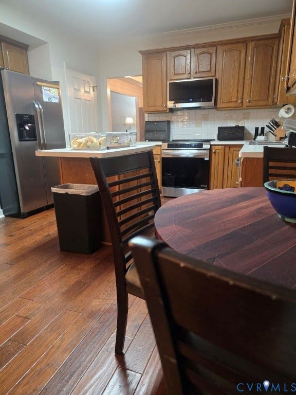 Wood-finish flooring throughout, wood cabinetry with brushed nickel hardware, stainless steel appliances, ceramic tile backsplash, and a breakfast bar