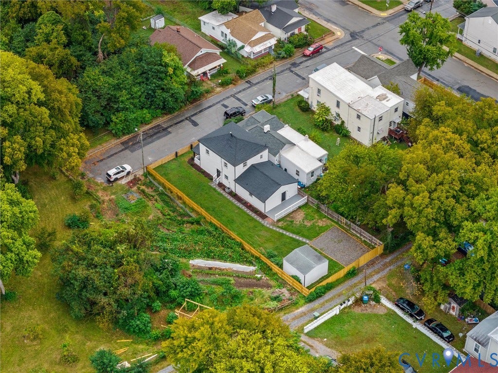 Aerial perspective of a property featuring a white residence with a dark roof, a fenced yard with green lawn, a detached storage shed, and a gravel parking area