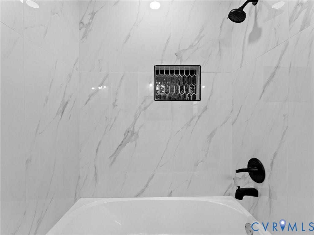 Bathtub with a matte black shower head and faucet, white tiled walls with subtle veining, and a recessed niche featuring hexagonal mosaic tile
