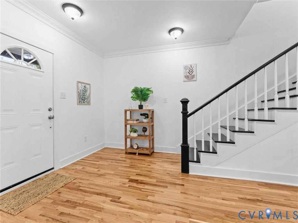 Hardwood flooring, white entry door with an arched transom, staircase with white risers and dark treads, white walls, and crown molding