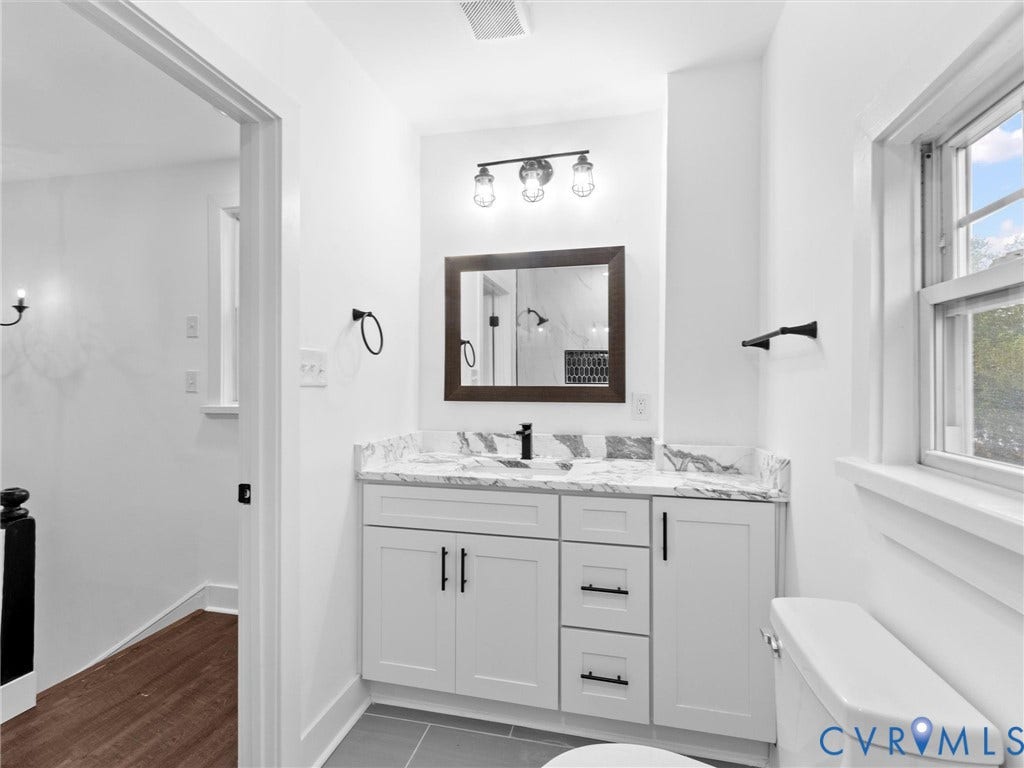 White vanity with black hardware, marble-finish countertop, dark-framed mirror, and industrial-style vanity lighting