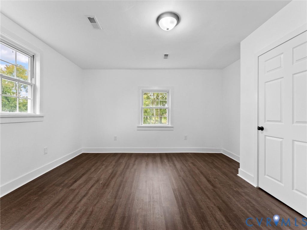 Wood-finish flooring, white walls, two double-hung windows with trim, flush mount ceiling light, and a paneled interior door with a black knob