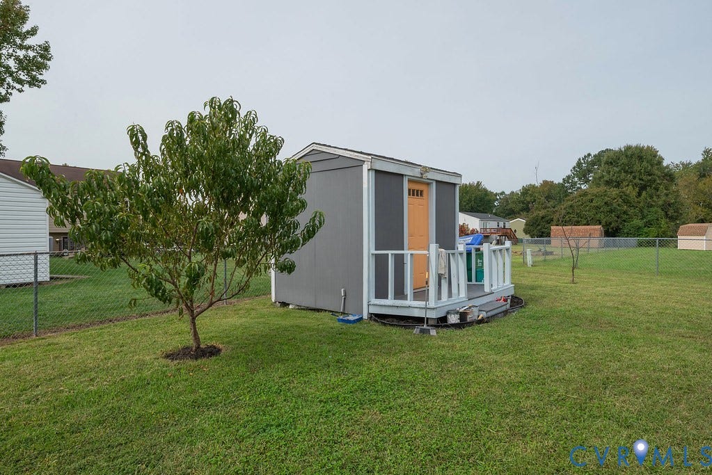 Detached storage shed with a built-in porch and an orange door, situated on a grassy lawn with a young tree