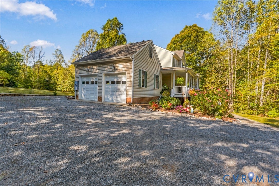 3647 Cedar Plains Road, Sandy Hook, Sandy Hook, VA 23153 Two-car garage with panel doors, gravel driveway, covered front porch with white railing, mature landscaping with flowering shrubs, and surrounding deciduous trees