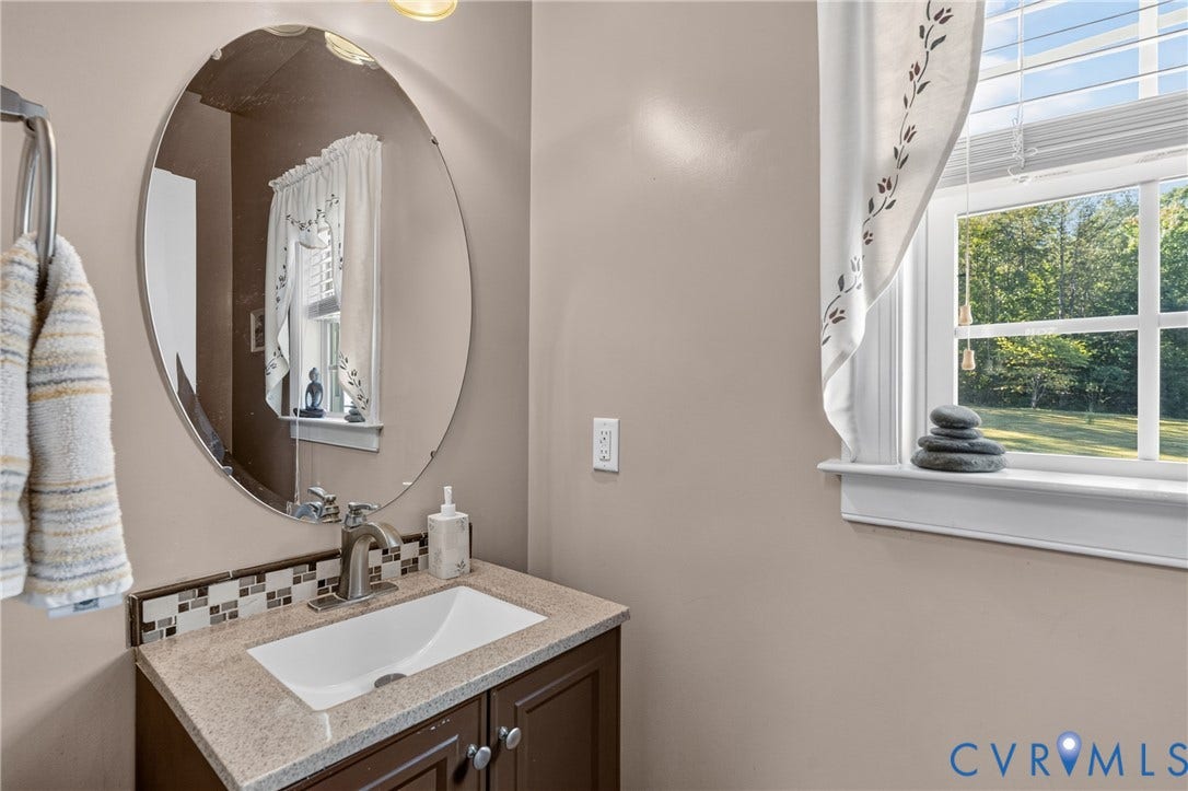 3647 Cedar Plains Road, Sandy Hook, Sandy Hook, VA 23153 Elegant powder room featuring a round mirror with brushed metal frame, floating vanity with stone countertop and rectangular vessel sink, mosaic tile backsplash accent, and abundant natural light from a large multi-pane window overlooking mature landscaping