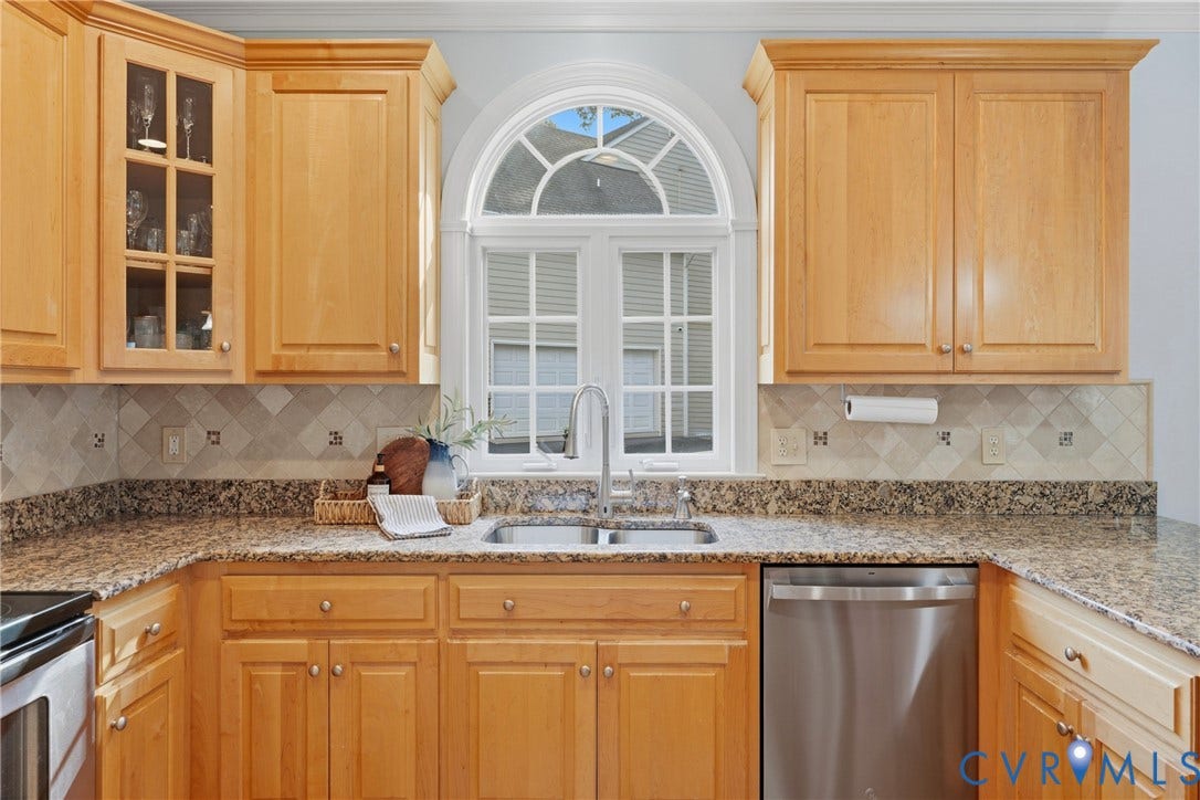 11813 Longfellow Drive, Bayhill Pointe, Midlothian, VA 23112 Kitchen featuring light wood cabinetry with glass-panel upper doors, stone-finish countertops, tile backsplash with decorative inserts, stainless steel double-basin sink with gooseneck faucet, and an arched window