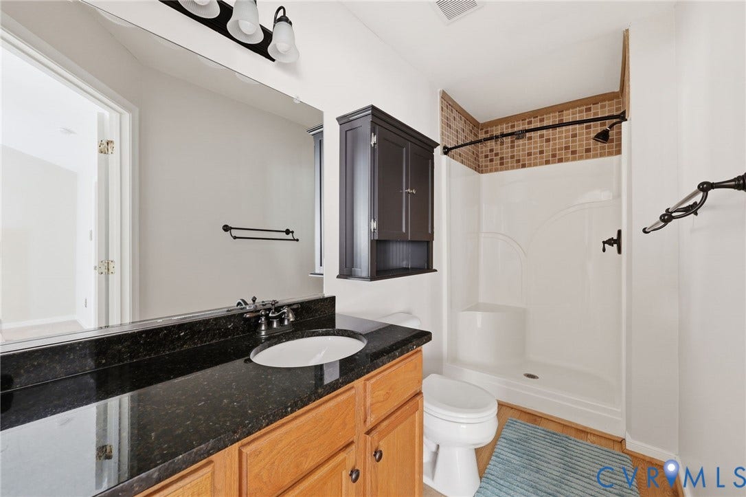 2803 Queensland Drive, #2803, Pemberton Oaks, Glen Allen, VA 23294 Dark stone-finish countertop with an integrated oval basin, natural wood-finish vanity cabinetry, wall-mounted dark cabinetry, and shower enclosure featuring tile-accented walls and built-in shelving