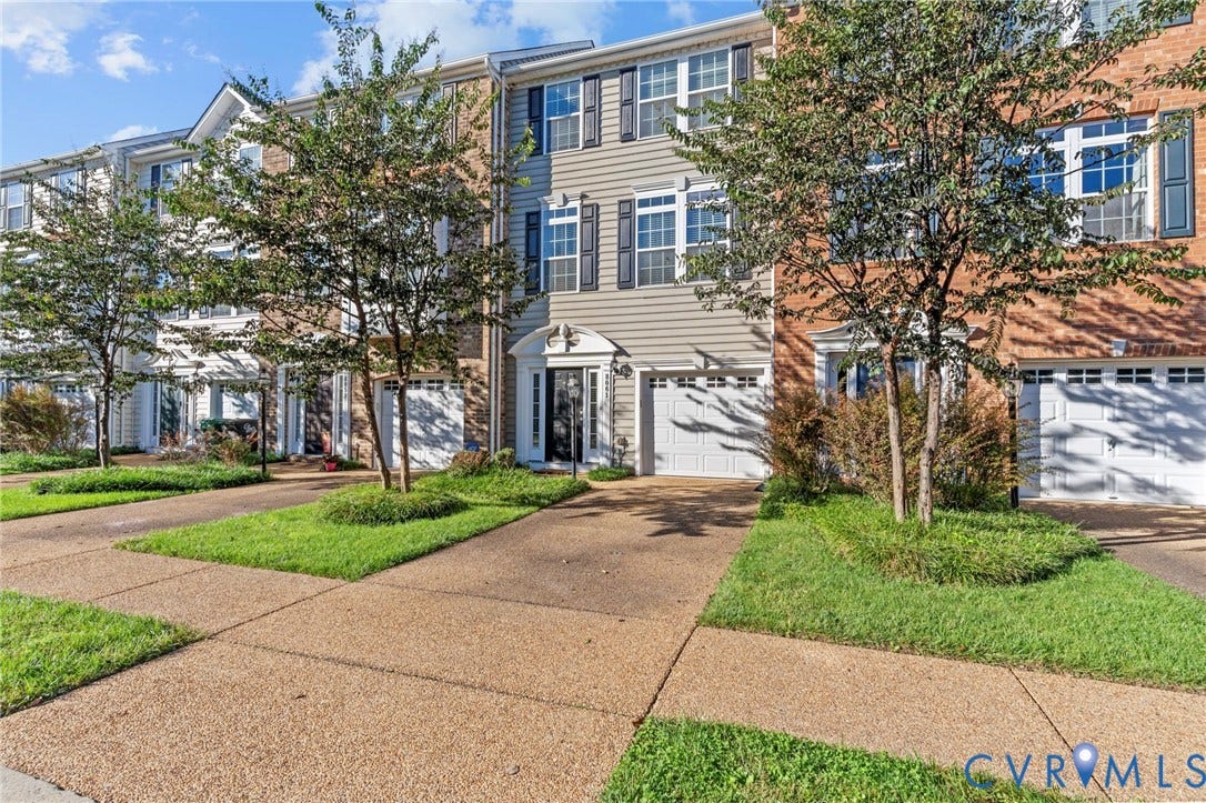 Three-story townhome with a light-toned facade and dark shutters, featuring a prominent garage door, front entry, and a well-maintained landscaping