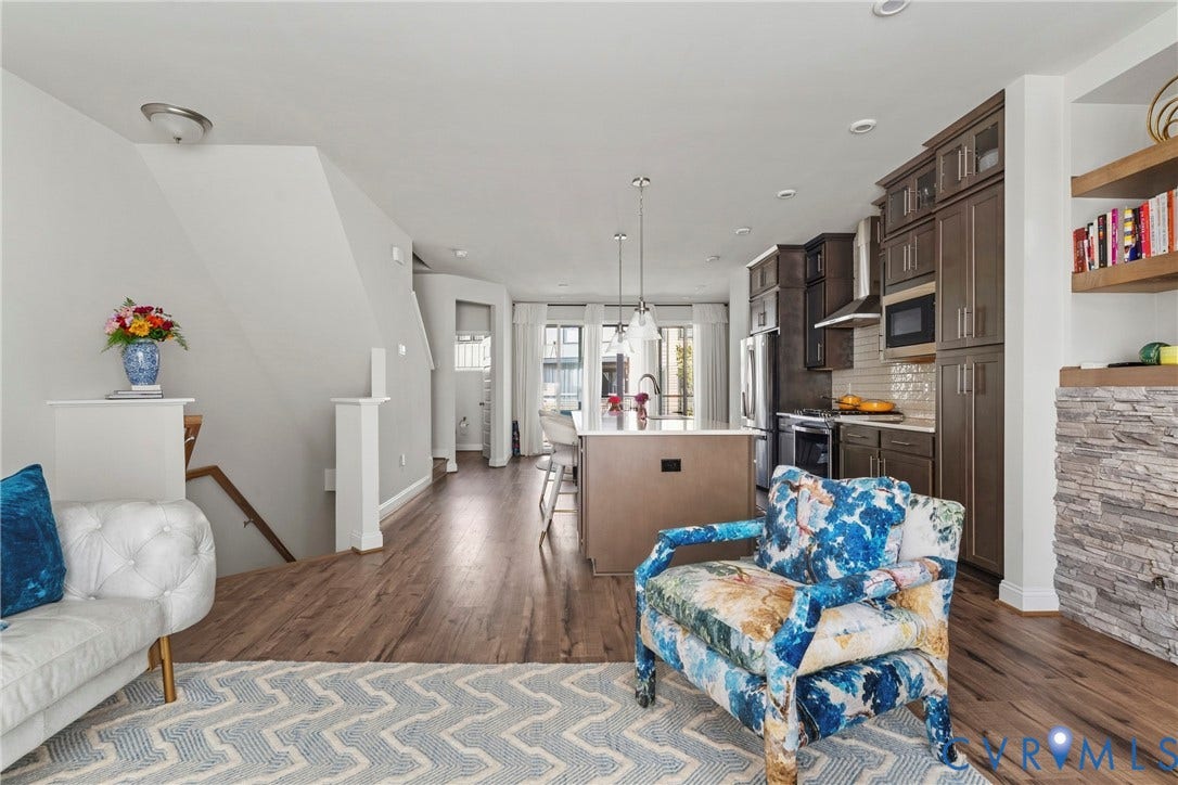 Open-concept living area featuring warm wood-finish flooring throughout, custom cabinetry with integrated shelving, a centered kitchen island with pendant lighting, and abundant natural light from multiple windows and glass doors
