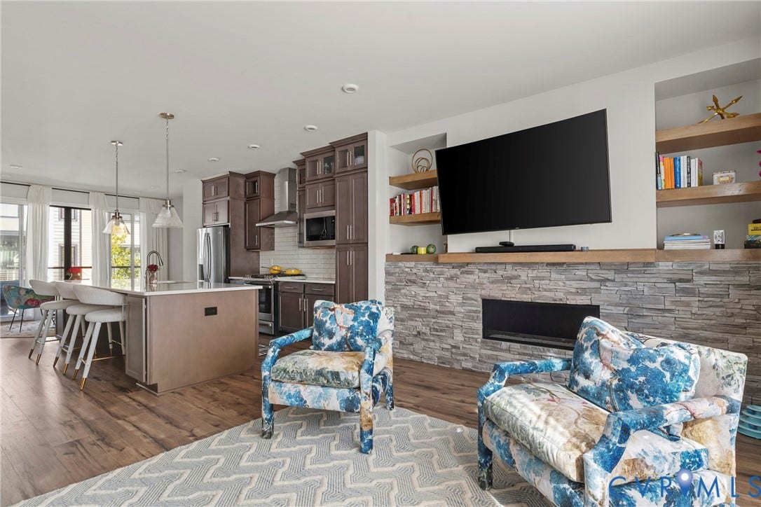 Open-concept living area featuring wood-finish flooring, a kitchen island with pendant lighting, dark wood cabinetry, and a fireplace with a stone surround and built-in shelving