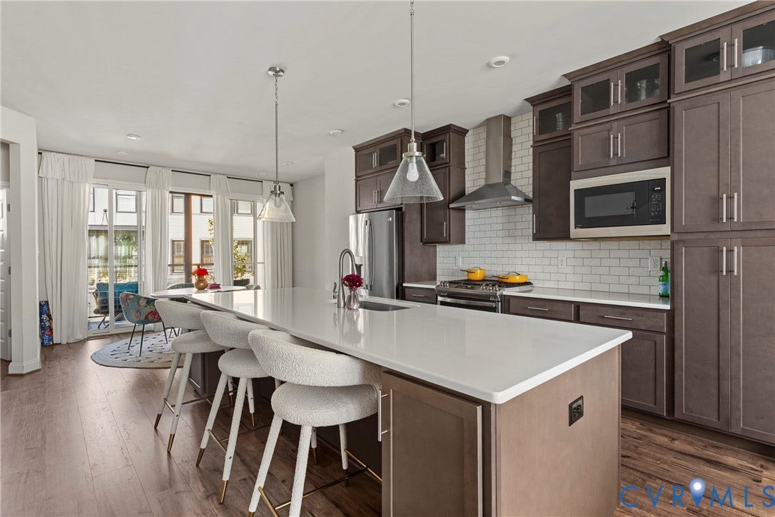 Kitchen featuring an expansive island with a pristine countertop, wood-finish flooring, ample cabinetry with glass-front uppers, a stainless steel range hood, and a subway tile backsplash
