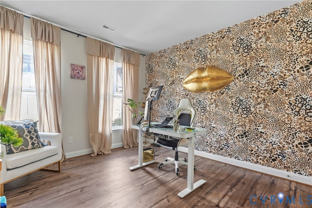 Bright, naturally lit home office featuring hardwood flooring, floor-to-ceiling textured accent wall with metallic wall sconce, expansive windows with neutral drapery, and dedicated workspace with built-in desk configuration