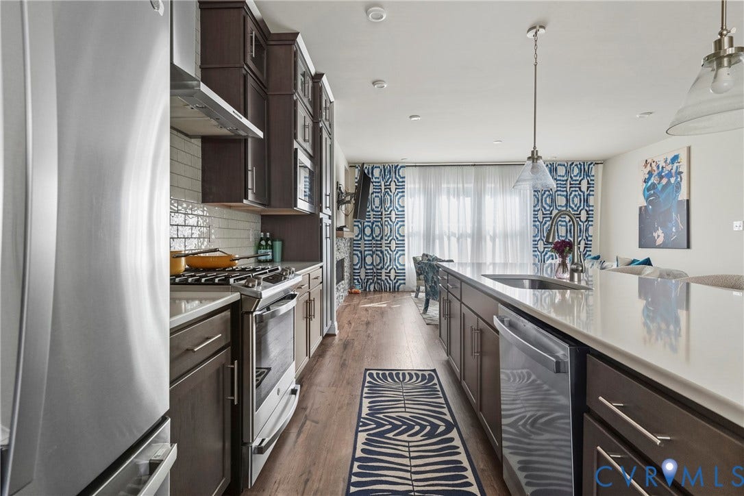 Kitchen featuring stainless steel appliances, dark wood-finish cabinetry, white subway tile backsplash, and an extended island with a single basin sink and pendant lighting