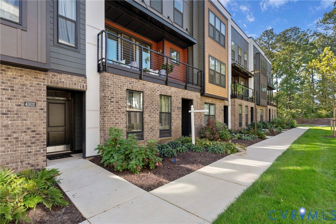 Modern townhome exterior featuring brick and horizontal siding, a private balcony with metal railings, a front entry door with transom window, and a concrete pathway