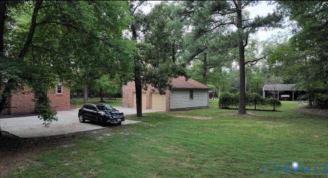 Expansive grassy yard with mature trees, a brick residence, detached garage with a light-colored door, a separate outbuilding, and a paved driveway