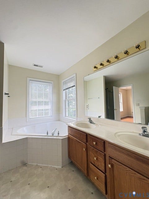 Dual vanity with integrated sinks, corner soaking tub, two windows with blinds, and a large mirror with overhead light fixture