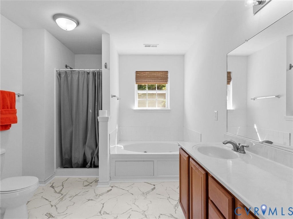 White tile flooring with grey veining, built-in bathtub, window with woven shade, and a wood-finish vanity with an integrated sink