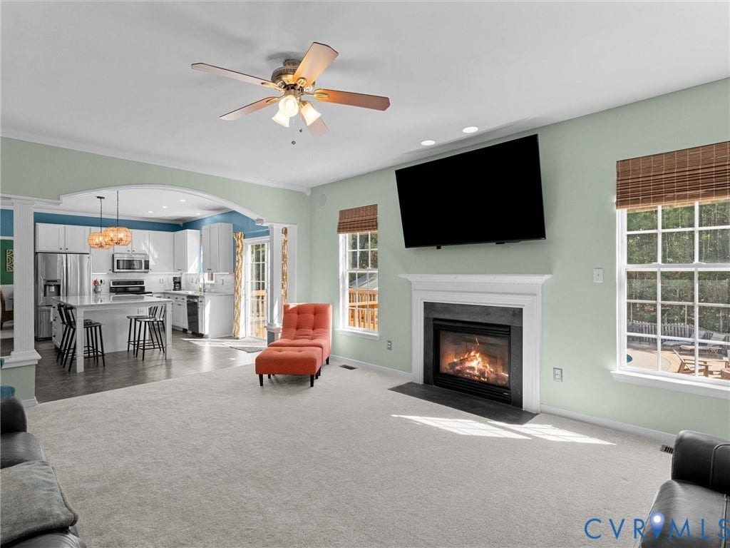 Spacious living area with a fireplace and white mantel, large windows with woven blinds, and a ceiling fan