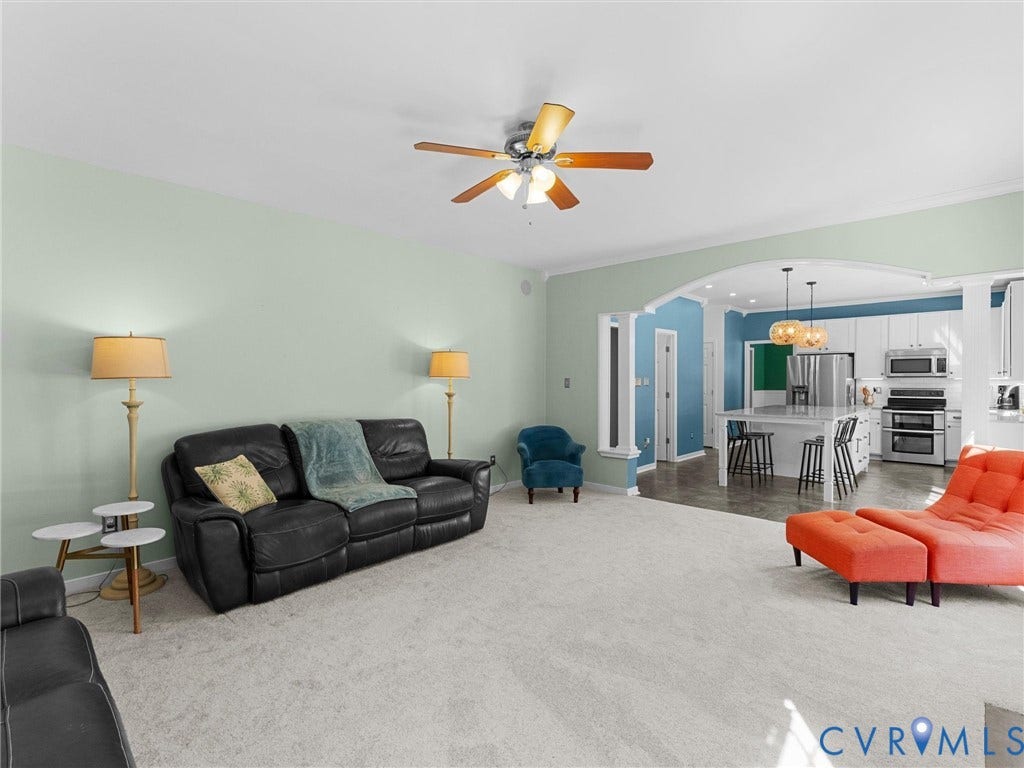 Spacious living area with light-colored carpeting, ceiling fan, and an arched entry leading to a kitchen with white cabinetry, stainless steel appliances, and a central island