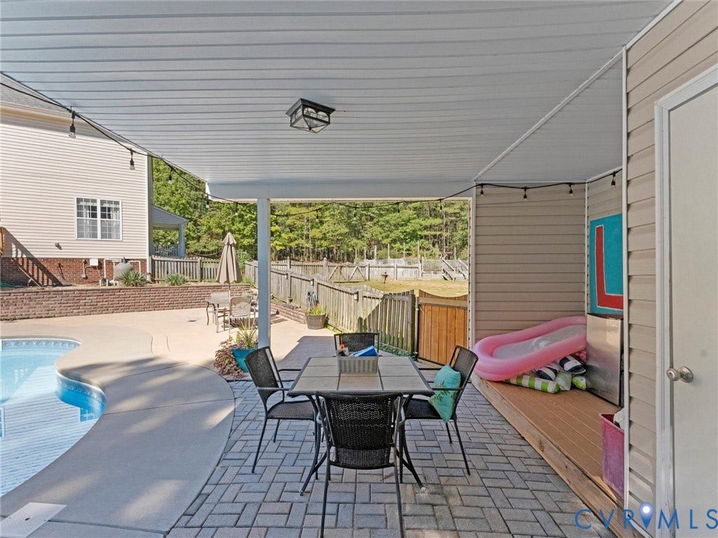 Covered patio with paver flooring, adjacent to a swimming pool with concrete decking, and a wood privacy fence
