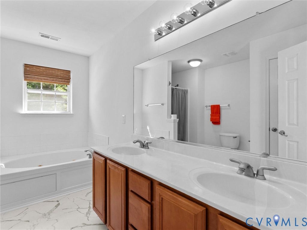 Dual sink vanity with wood-finish cabinetry, a large wall-mounted mirror, and a built-in soaking tub with a window