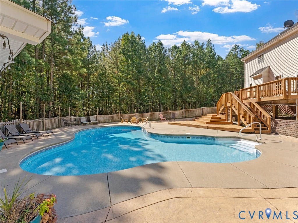 Curved in-ground pool with concrete surround, multi-level wood deck, and partial brick retaining wall