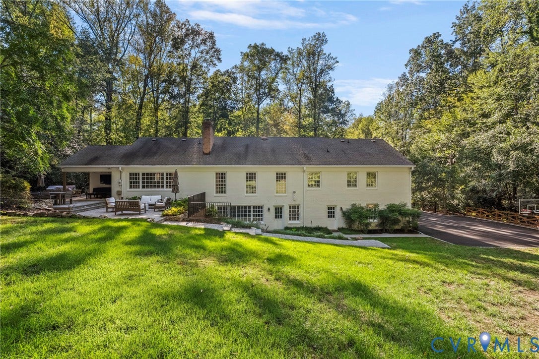 Expansive green lawn with a white brick exterior, a covered outdoor patio, and a paved driveway with a basketball hoop