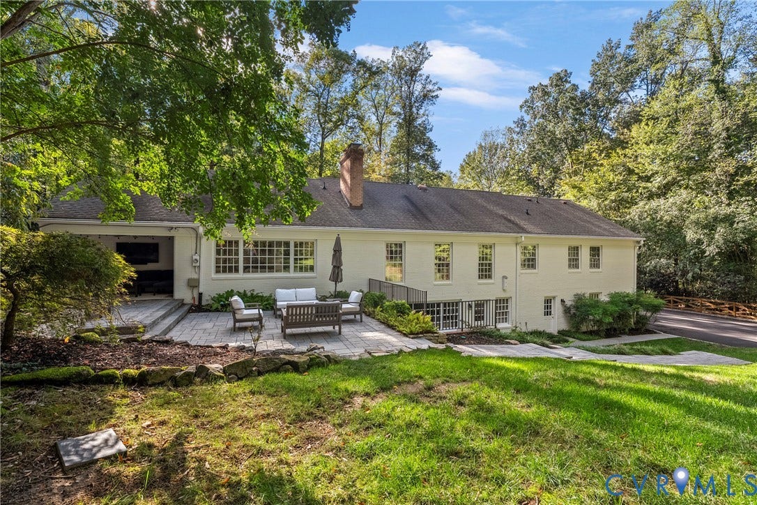 Expansive rear exterior featuring a paved patio, covered outdoor living space, multiple window installations, and a brick chimney
