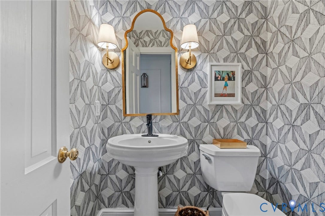 Pedestal sink with black faucet, brass wall sconces, decorative wall covering, and a toilet