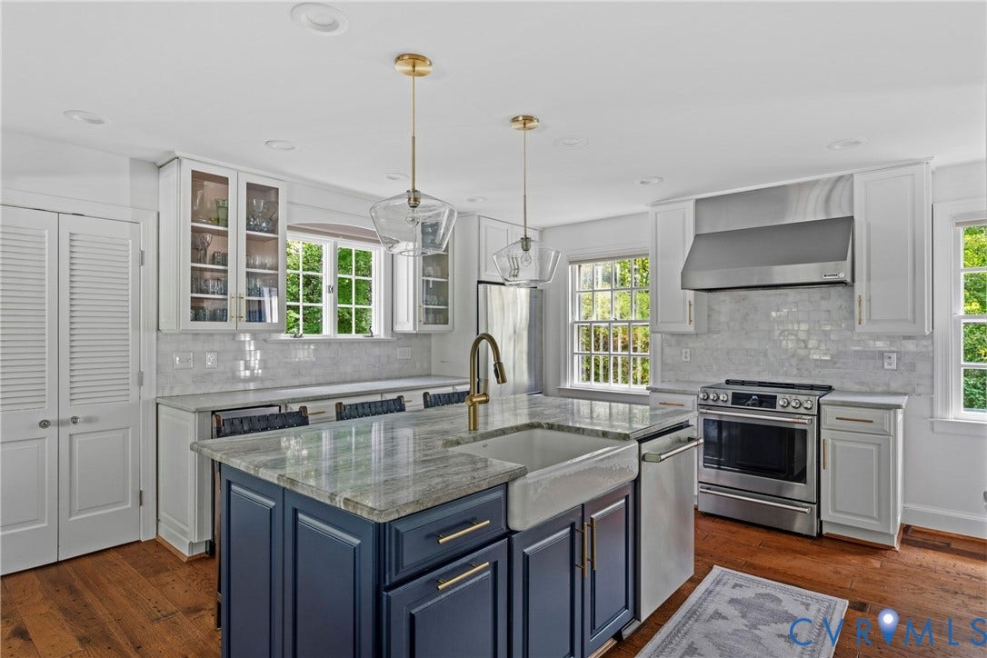 Expansive kitchen island with a farmhouse sink, stainless steel appliances, white shaker cabinetry, gold-finish hardware, and wood-finish flooring