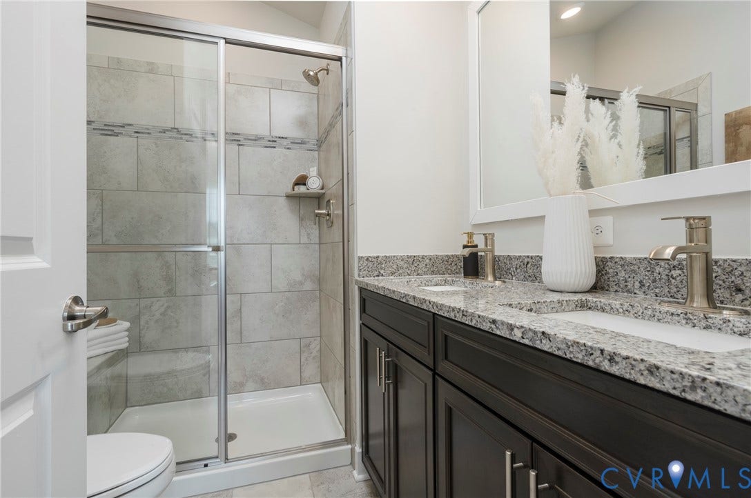 Dual vanity with granite countertops, dark wood-finish cabinetry, brushed nickel fixtures, and a glass-enclosed shower with tiled walls and a mosaic accent band