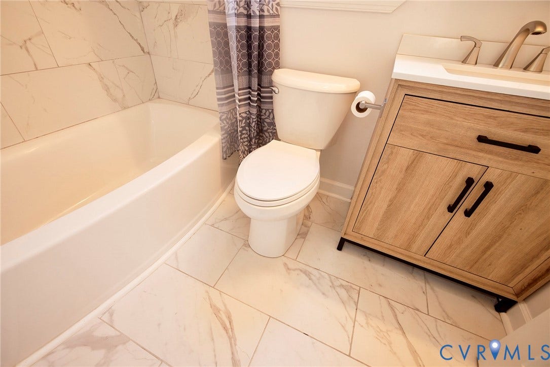 2421 Buena Vista Boulevard, Chester, VA 23831 White rectangular bathtub with tiled surround, wood-finish vanity with drawer and cabinet, brushed nickel faucet, and marble-look floor tiles