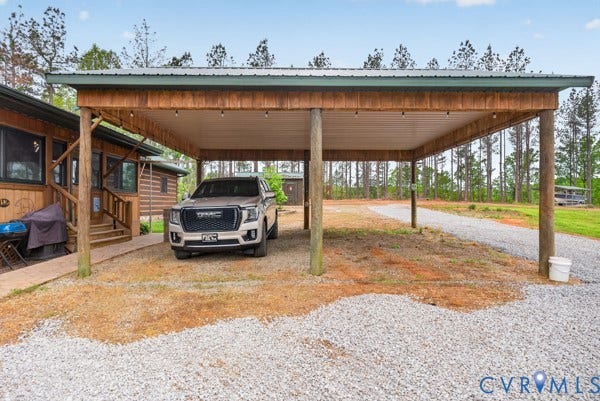 Expansive gravel driveway and carport with wood posts, wood-finish ceiling, and string lighting, beside a wood-sided structure