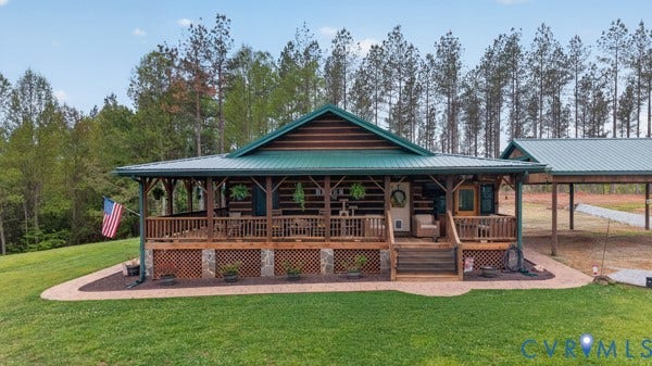 Log cabin exterior with a green metal roof, a full-length covered wooden porch, stone accents, and a paved walkway