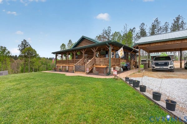 Log cabin exterior with a covered front porch, an expansive lawn, a gravel driveway, and a detached covered carport