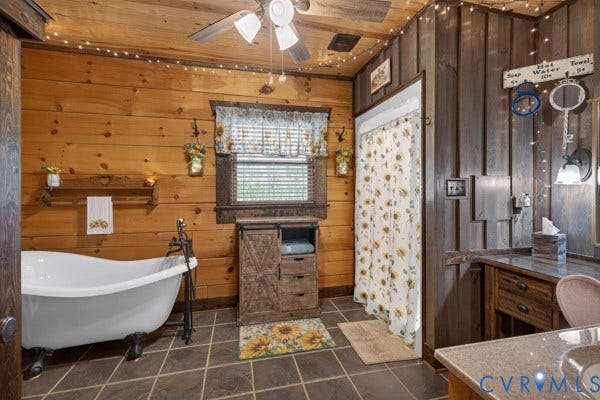 Freestanding soaking tub with floor-mounted fixtures, wood-paneled walls, tile flooring, vanity with countertop, and wood-finish ceiling fan