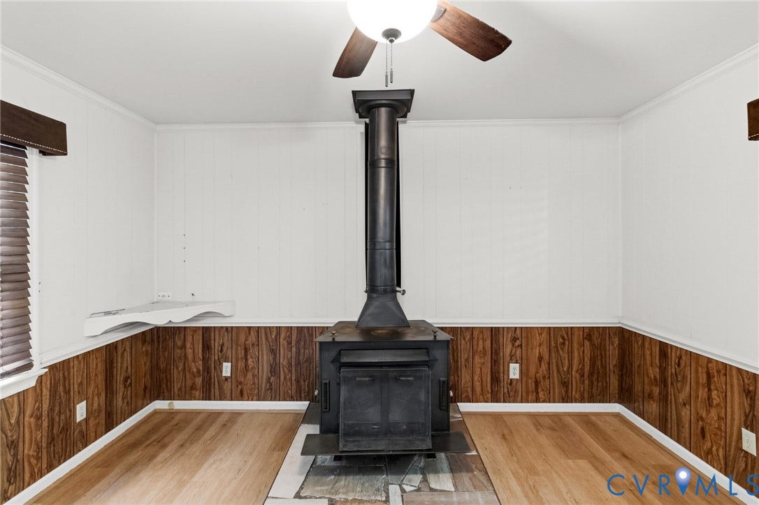 7847 Beaver Drive, All Others Area 20, Gloucester, VA 23061 Wood-look flooring, ceiling fan with integrated lighting, wood-paneled wainscoting, and a central wood-burning stove with a stone hearth
