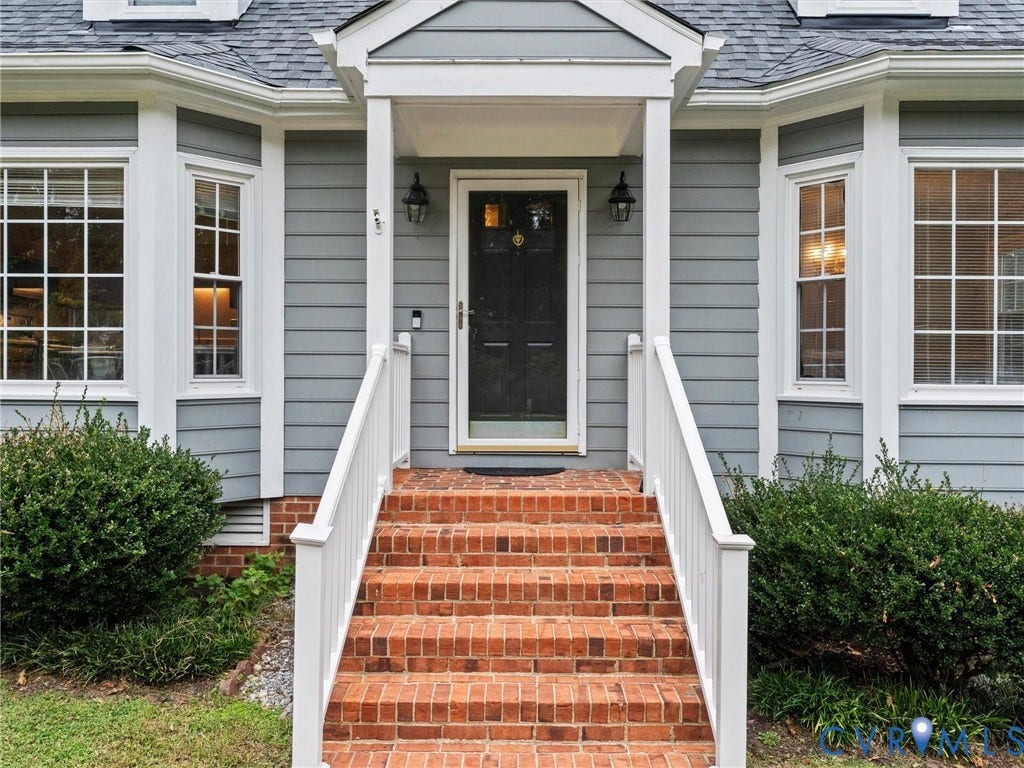 Exterior entrance featuring horizontal lap siding, multi-pane bay windows, a covered portico with white railings, and brick masonry steps