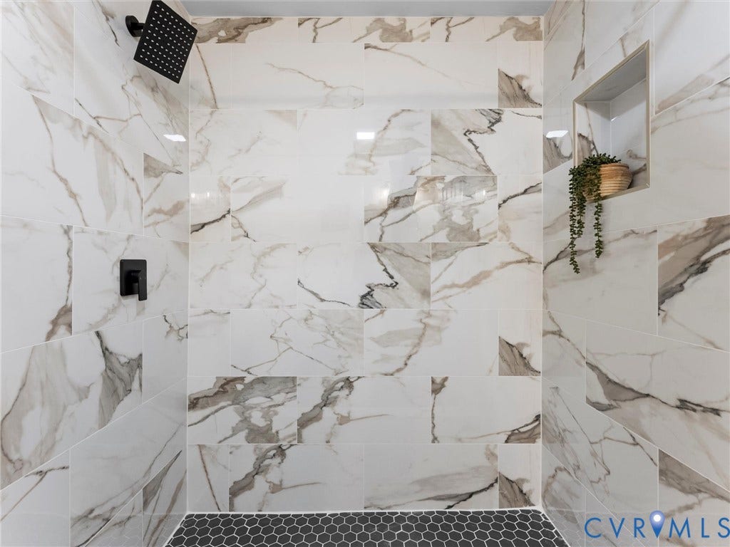 Shower with large format white and gray veined tile, matte black shower head and controls, black hexagonal floor tile, and a built-in shower niche