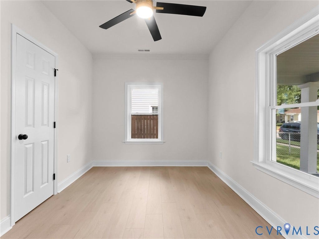 Light wood-finish flooring, white walls, two white-trimmed windows, a white paneled door with black hardware, and a ceiling fan